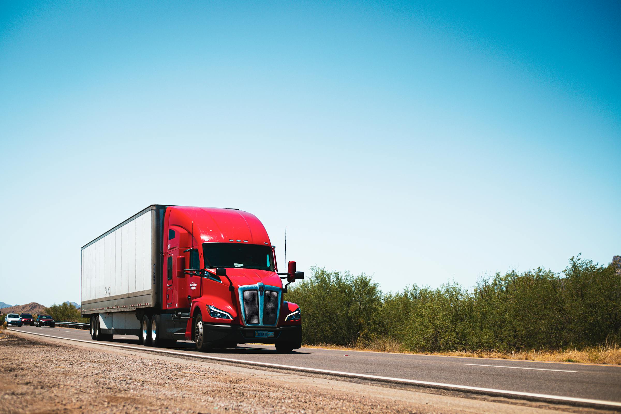 A red American semi-truck hauling a dry-van trailer on an open highway under a clear blue sky