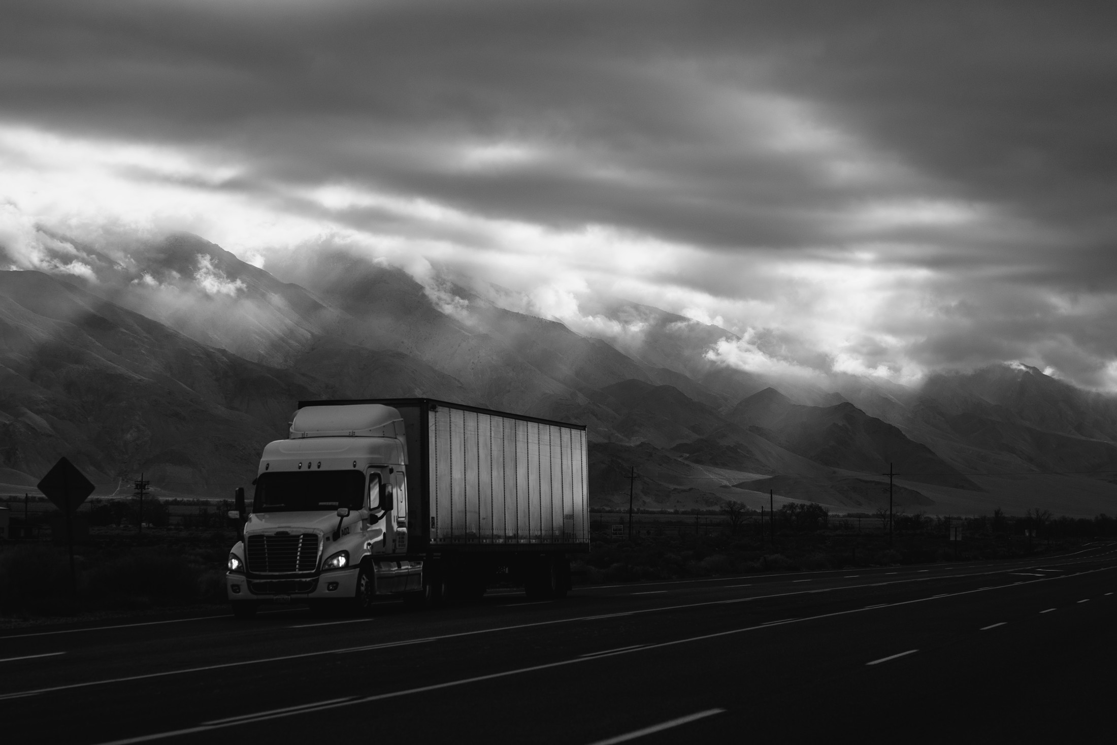 A semi-truck driving a mountain highway beneath a dramatic, cloudy sky